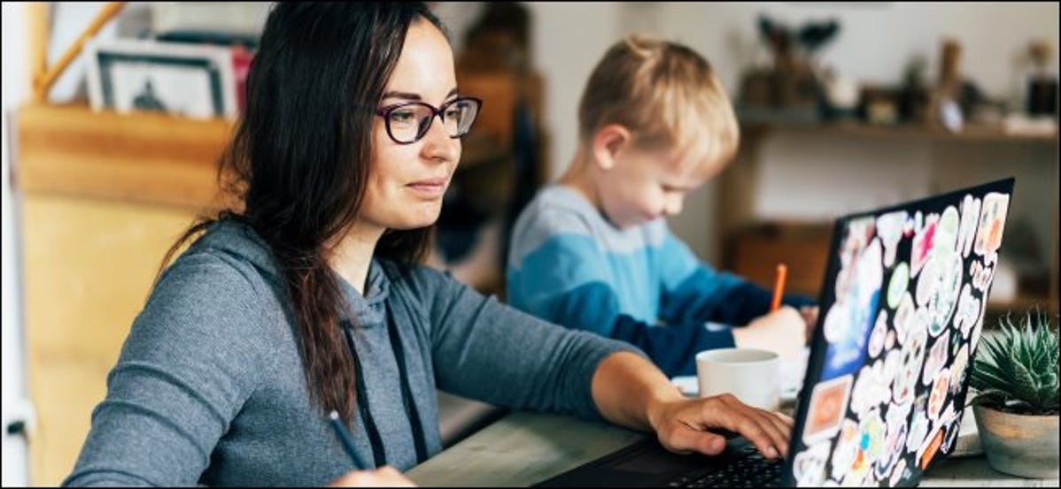Seorang wanita bekerja pada laptop dengan seorang anak duduk di sebelah gambarnya.