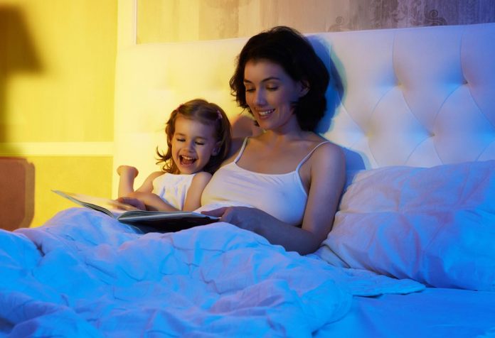 Seorang ibu dan anak perempuannya membaca buku bersama di tempat tidur