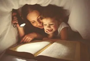 Seorang ibu dan anak perempuannya membaca buku di bawah senter di malam hari